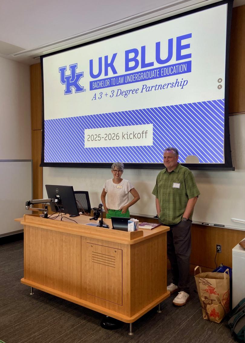Two people stand at a podium in a classroom with a large screen behind them displaying "UK BLUE 2025-2026 kickoff: Bachelor to Law Undergraduate Education.